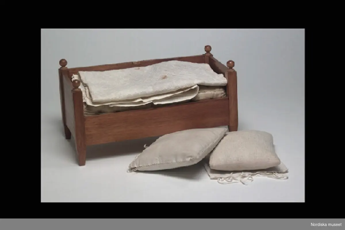 Inventering Sesam 1996-1999:
Säng med sängkläder
a) säng L 32,5 cm, B 16 cm, H 17 cm
Tillverkad av brunbetsad sockerkist (=ceder). Gavlar och ena långsidan lika höga, den andra långsidan låg, hörnstolparna krönta med knopp. Benen avsmalnande nedåt. 
b) madrass L 28 cm, B 14 cm, H 7 cm.
Tjock madrass av vit tuskaft i bomull (?) stoppad med tagel.
c) örngott 14,5 x 13,5 cm
Örngott av vitt linne, ihopsydd med dekorsöm på tre sidor, öppen och med knytband på den fjärde sidan. 
d) kudde 12,5 x 14 cm
Av vit bomull, rosa band runt kanterna, stoppad med fjäder.
e) Kudde
Lika som d).
f) Örngott 14,5 x 15,5 cm
Något större, i övrigt lika som c). 
g) Lakan L 41,5 cm, B 31 cm. 
Av tunt linne med spets i ena kortändan.
h) Täcke L 35 cm, B 26 cm,
Av bomullstyg (enligt tidigare katalogkort "skärt", nu blekt) täckt av vit tyll som är broderad med vit tråd.
Bilaga =  "Har haft till egarinnor fru Louise Sterky, född Sterky, och Carin Sterky, född Sterky, döttrar till grosshandlare Fredrik Sterky, Stockholm". (framgår ej om bilagan gäller enbart dockorna i gåvan eller samtliga föremål)
Leif Wallin 1996