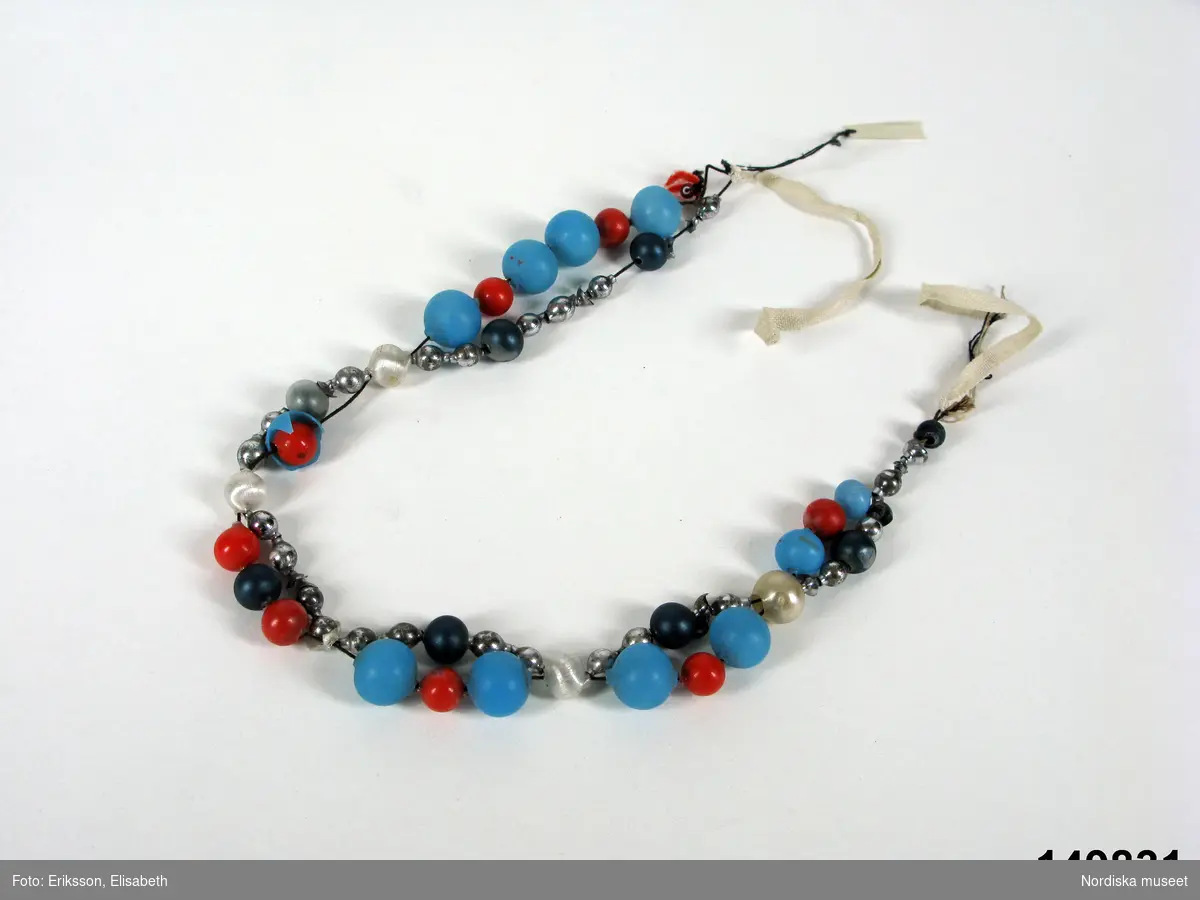 Halsband av blå, röda , vita och vitmetallblanka glaspärlor trädda i 2 rader som går ihop på jämna mellanrum.
Anm. Några pärlor skadade.
Har ingått i en brudutstyrsel.

Berit Eldvik maj 2006