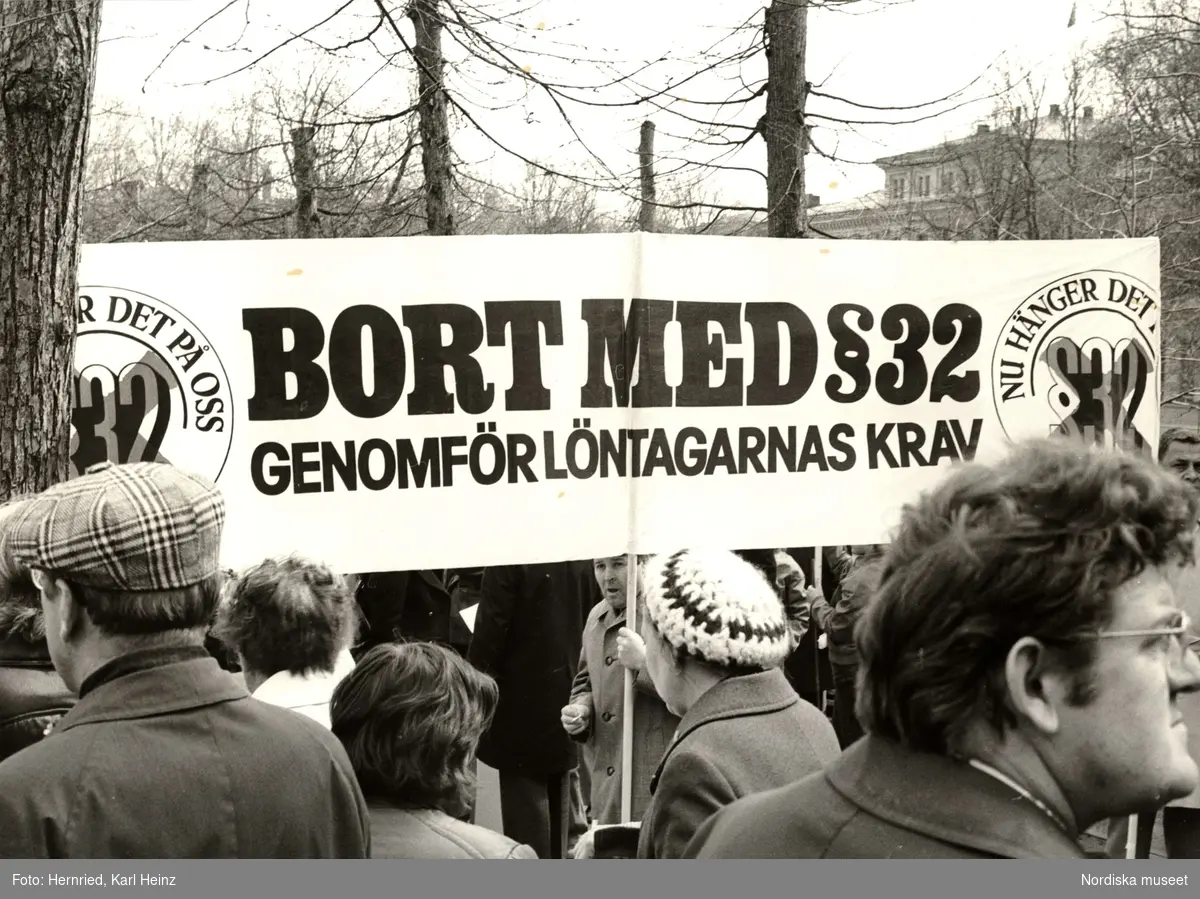 Demonstration 1:a maj. Plakat med texten "NU HÄNGER DET PÅ OSS. BORT MED §32. GENOMFÖR LÖNTAGARNAS KRAV