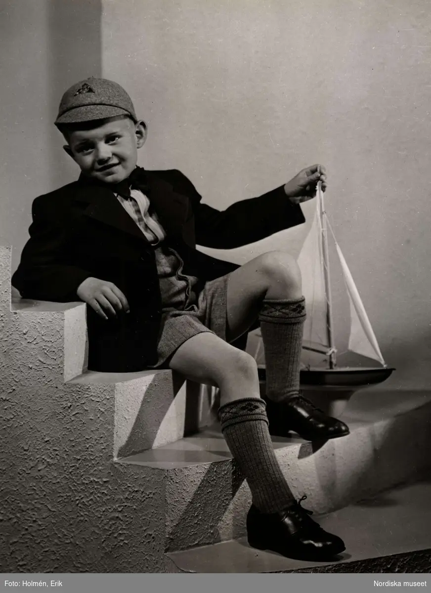 Barnkläder visas på Nordiska Kompaniet 1941. Pojke sitter i trappa iklädd kavaj och kortbyxor, knästrumpor, knytskor och keps. Han håller en segelbåt med ena handen.