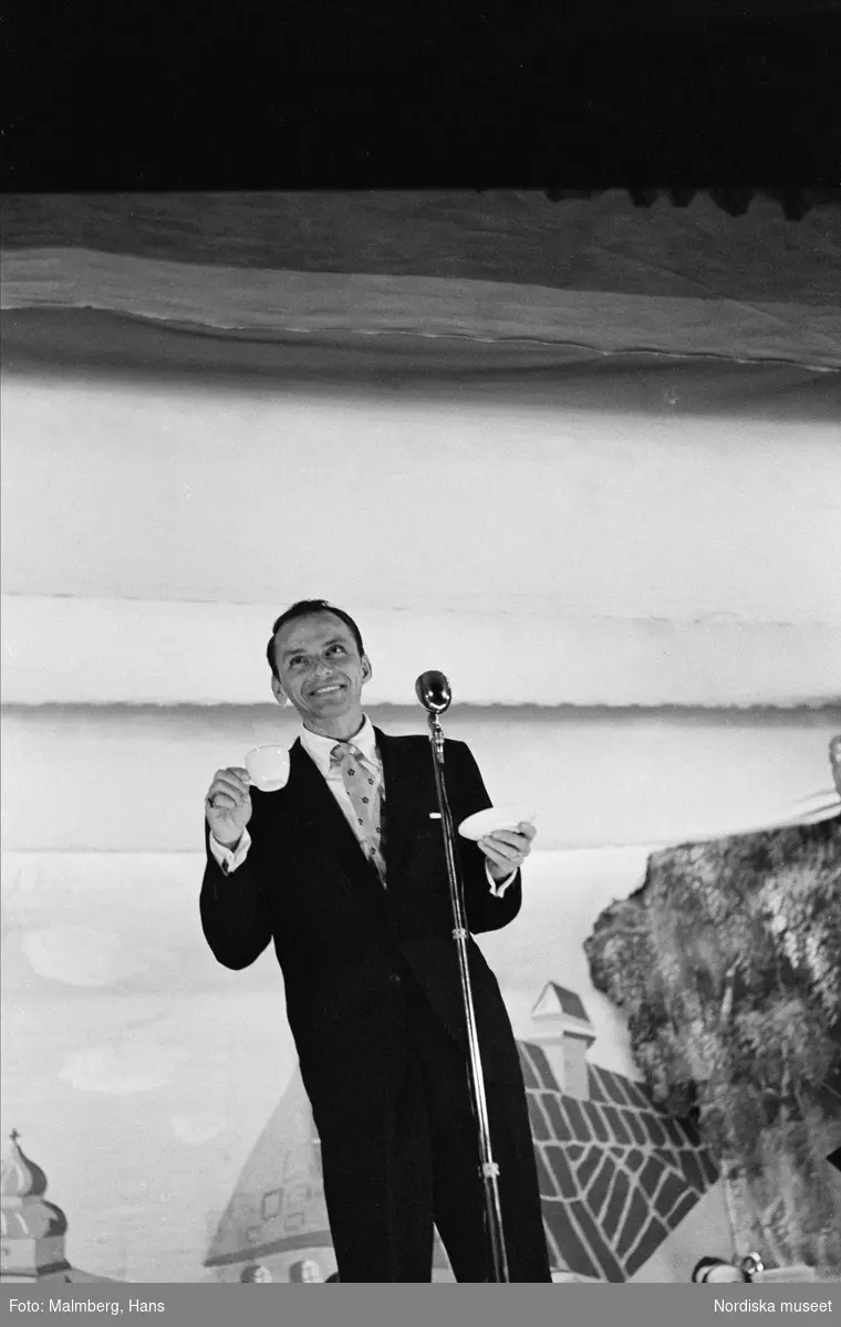Frank Sinatra på turné i Sverige. Finspångs Folkets park. 
Sinatra framför mikrofonen på scenen, med en kaffekopp och ett kaffefat i vardera handen. 