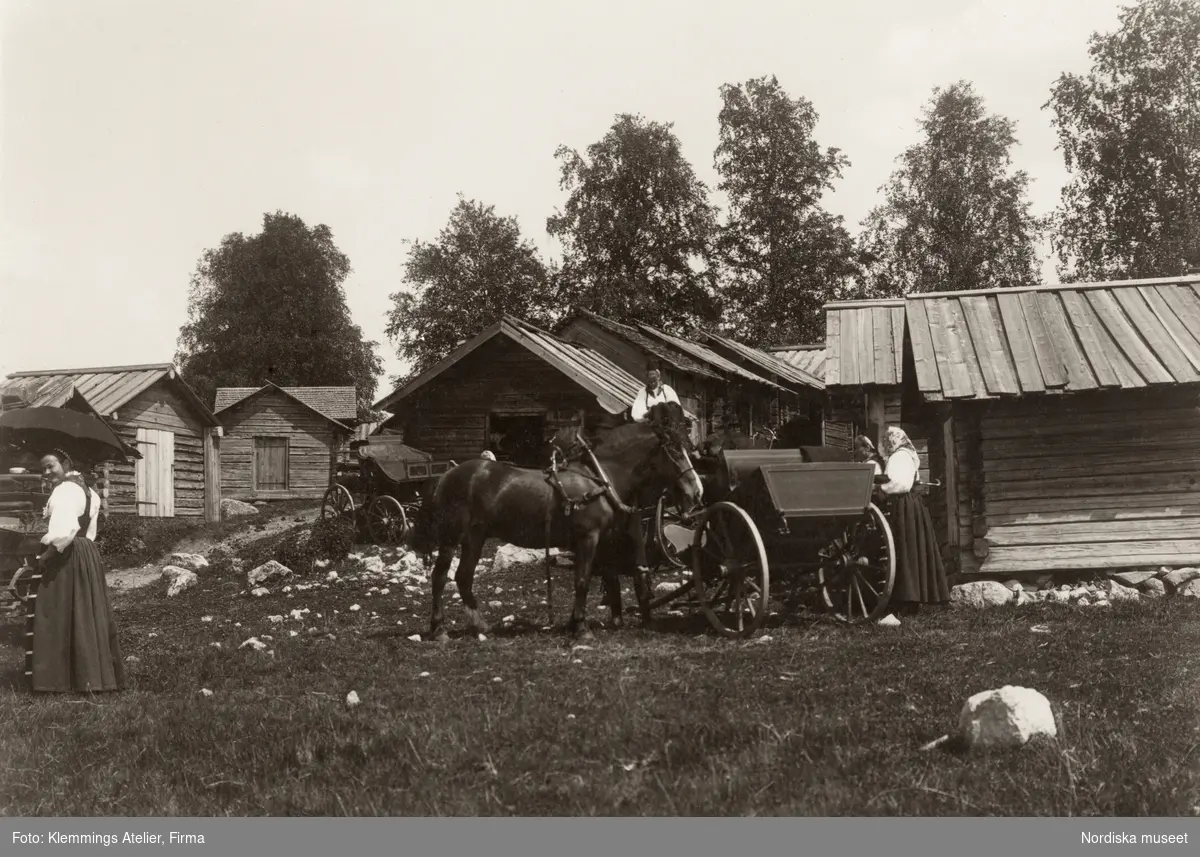 Kyrkstallarna vid Rättviks kyrka i Dalarna. Framför stallarna står vagnar, en häst och några kvinnor i högtidsdräkt.