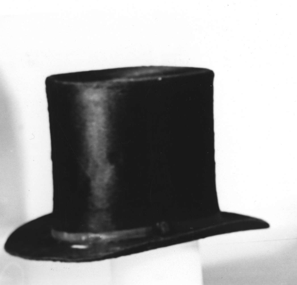 Hög hatt av svart långhårig felb. Runt brättet ripsband med svartlackerat spänne. Fodrad med veckad vit papp med påklistrad etikett med text: N G Ögren. Till hatten hör en ask (UM08649b).