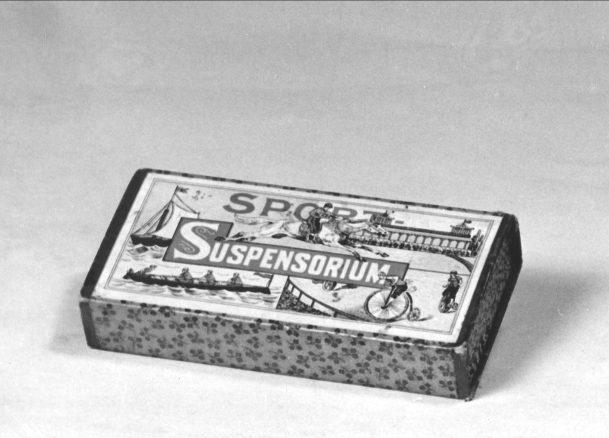 Suspensoar i turkos ask av papp med gulddekor och kanter. Etikett i grågrönt, svart, vitt och rött med text "Sport-Suspensorium". Med blyerts står "2:-". Suspensoaren är tillverkad av vit och ljusblå textil.