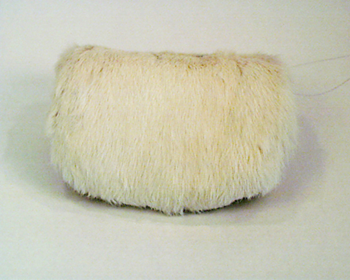 Muff med vitt kaninskinn på framsidan och grå sammet på baksidan, ficka med blixtlås på baksidan. Fodrad med vit bomullslärft, stoppad. Ingen hängband.