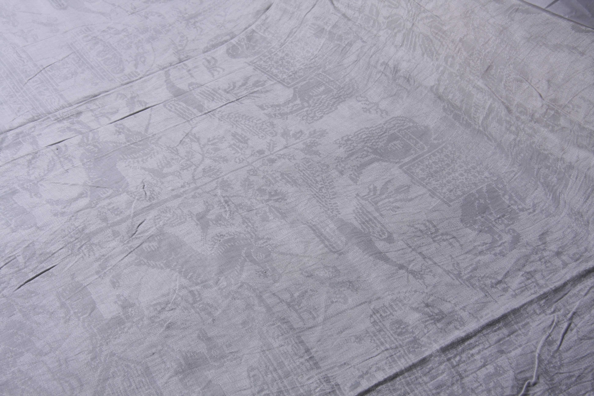 Hvit lindamask med mønster: Enhjørninger flankerer fontene stående på bakbenene med liggende hjerter og andre dyr omkring. Damer til hest med hatt på hodet. Duken har smal fall på kortsidene.