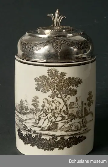 Svagt, uppåt avsmalnande kärl med raka sidor och en hänkel.
Vit (beige) med brun-svart tryckt dekor - ett landskapsmotiv med en sittande man och kvinna samt en hund och två får.
I bakgrunden är det träd och hus. I förgrunden är det låg vegetation. Kärlet kan vara tillverkat i Norge.
Kärlet har ett lock av silver fäst i hänkeln.
I locket är ett förgyllt silvermynt från 1733 med Christian VI i profil infattat. Han var kung över Danmark och Norge.
Locket har ingraverade blommor och girlanger av blad samt monogram: "OBS" och "AEP".
På baksidan av den detalj som sammanbinder lock och kärl står det: "Vir 7027" (?).
Locket har fyra silverstämplar i från vilka följande kan utläsas:
Tillverkat i Bergen, Norge av Andreas Kierumsgaard Saebyes änka, under perioden 23/9 - 22/10 (enligt månadsstämpel; vågen) 1797.
Föremålet har Mathias Pettersens kontrollantstämpel (wardeinsstämpel). Han var kontrollant 1790 - 1812.
Kärlets mynningskant är mycket söndrigt. Kärlet har sprickor.

Ur handskrivna katalogen 1957-1958:
Krus av porslin m. lock av silver
Bottendiam.: 9,4. H: (med lock) 14,1. Vitt porslin med landskapscen i brunsvart. Locket ornerat. Sprucken och lagad.

Lappkatalog: 62