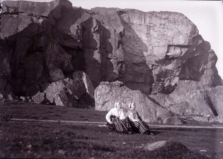 Enligt text som medföljde bilden: "Karin och Lisa. 2 kvinnor klädda i folkdräkt sitter på en äng framför en bergvägg."