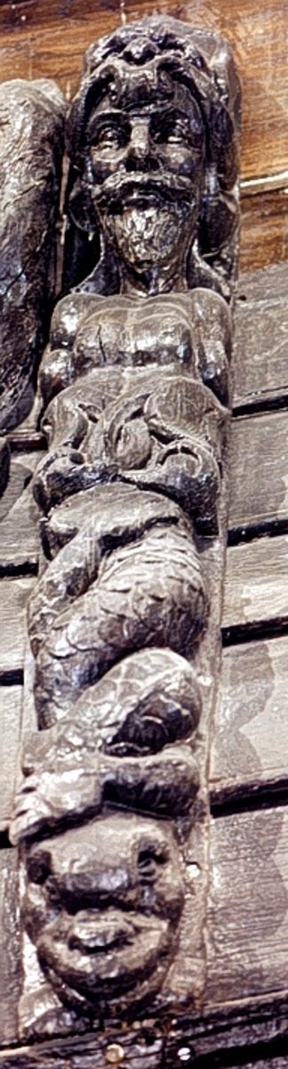 Triton med kraftig och bred näsa, stora mustascher och spetsigt skägg. På huvudet en hjälm med en djurmaskaron, eventuellt föreställande ett lejonhuvud. Hjälmen har en profilerad framkant prydd med en rullverksvolut.

Armarna har ersatts med stora dekorativa bladslingor. Skulpturens mittparti består av två krysslagda bladslingor med upprullade och förtjockade ändar. Tritonens fiskstjärt slingrar sig kring stjärten till en fisk eller en delfin vars huvud är synligt vid skulpturens fotända.

Bakstycket är kraftigt och snidat i ett med skulpturen. Baksidan är försedd med hak eller urtag. Skulpturens profil är svagt S-formad.
Skulpturen är mycket välbevarad.

Text in English: Triton with thick, broad nose, large moustache and pointed beard. The figure wears a helmet with an animal mask, probably a lion''s head. The helmet has an elegantly profiled front edge, decorated with scroll volutes. The arms are replaced by leafy ornamentation. The middle section is dominated by two crossed leafy vines with rolled-up, thickened ends. The fish tail is entwined with the tail of a fish or dolphin which can be seen at the sculpture''s foot end.

The figure is carved in one piece with a robust backboard. The back side of the backboard is carved with hollowed out grooves. The profile is slightly S-shaped.
The sculpture is very well preserved.