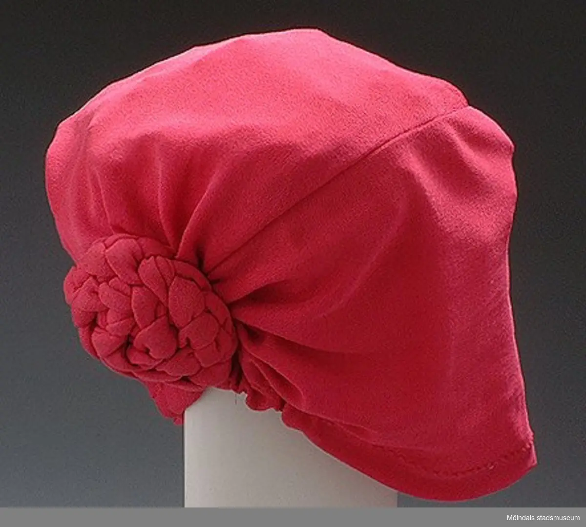 Rosa hatt/mössa med en knut bak på.Det finns också tillhörande blus, kjol och kavaj: invnr. MM03346:1, MM03346:2, MM03346:3.Måtten:Höjd 200 mm, diam. 200 mm. 