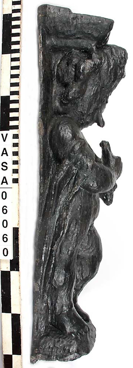 Skulptur föreställande en barnfigur som spelar på en sinka (ett äldre blåsinstrument med grepphål som blockflöjten men ett munstycke som liknar trumpetens).
Huvudet är vridet något åt höger. Kroppstyngden vilar på höger ben medan vänster ben är böjt. Instrumentet hålls med båda händerna. Skulpturen förefaller delvis draperad i en mantel eller dylikt som skymtar vid hans högra sida. Ovanför hjässan syns en något snedställd äggstavslist. Figuren står på ett minimalt underlag som troligen avser att föreställa ett stycke naturlig mark. Skulpturens baksidan är slät.
Skulpturen är kraftigt sliten.

Text in English: A sculpture of a child playing a sinka.
The weight of the body rests on the right leg while the left is bent and the head is turned slightly to the right.. The instrument is held in both hands. The figure seems to be partly draped in a cloak or the like, which conceals his right side. Just above the head the back board comes forward at right angles and the front of the board is decorated with an egg  mould design. The sculpture stands on a small flat plinth. The back of the sculpture is smooth.
The sculpture is very worn.