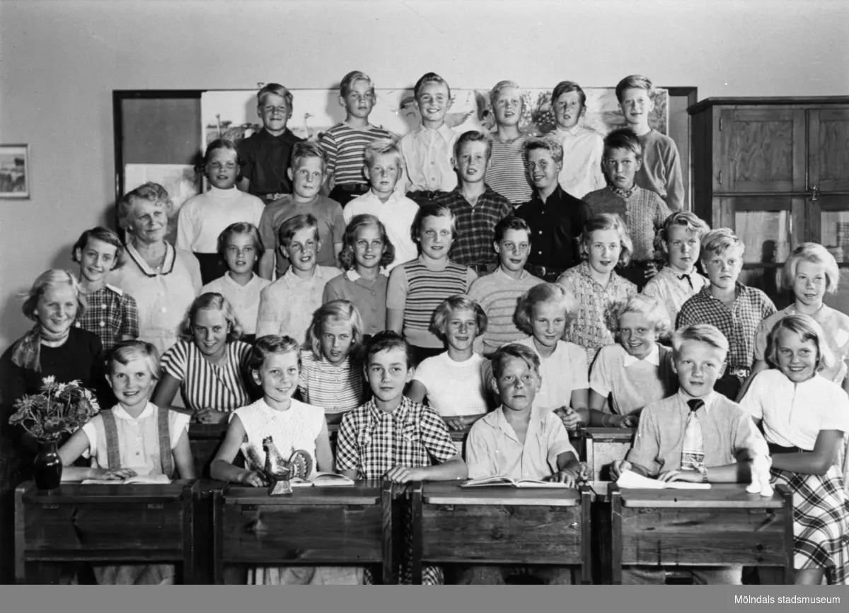 Flickor och pojkar stående i klassrum på Kvarnbyskolan i Mölndal år 1952. De två främsta raderna sitter i bänkar. Med på fotot är givaren Margit Hellström (f. Willumsen).