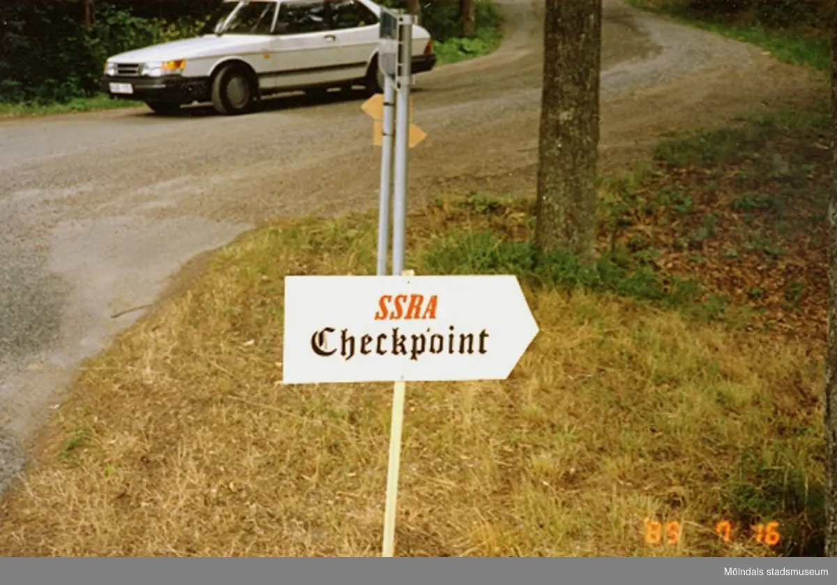 En skylt med texten "SSRA Checkpoint" som står uppställd i Gunneboskogen. I bakgrunden ser man en vit Saab som kör på en grusväg.
