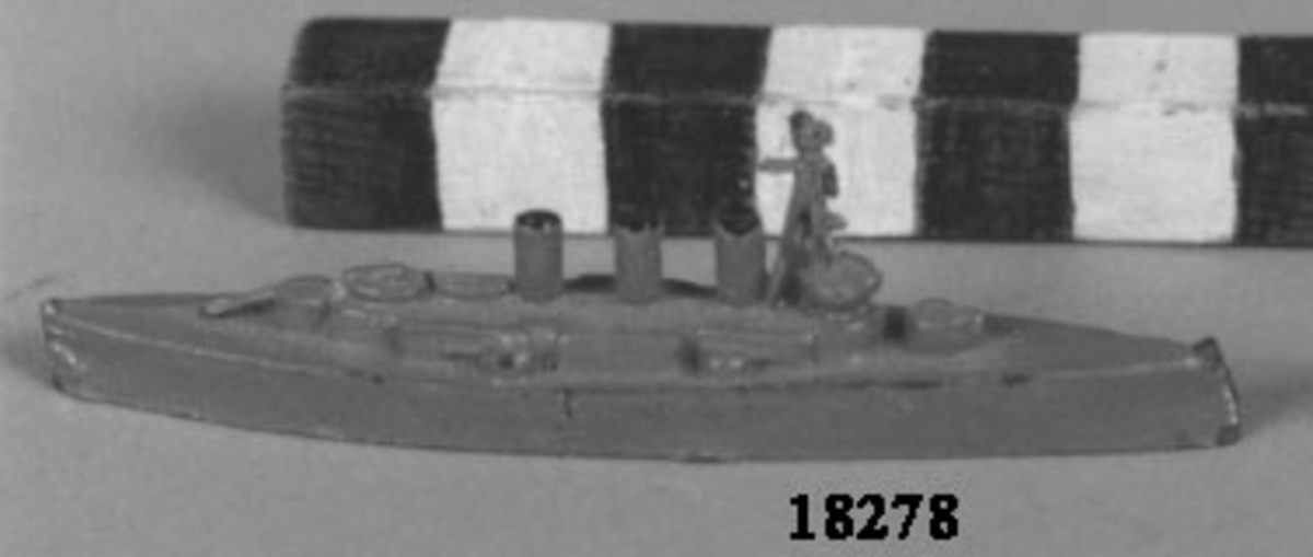 Fartygsmodell i form av pansarskeppet Oscar II gjuten av metall i ett stycke, målad i grått. Spetsig för och akter. Plan botten. Artilleripjäser i för, akter och sidorna. Mast samt tre skorstenar. I botten skrivet fartygets namn.