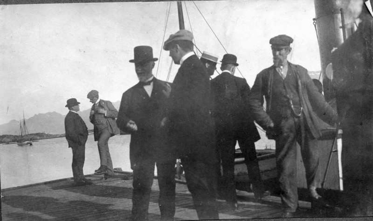 Fra kaia i Melbu havn. Flere menn i samtale. Ved kai ligger en fiskedamper med en dory om bord. Mannen midt i bildet med skyggelue kan være Jens Frederiksen.