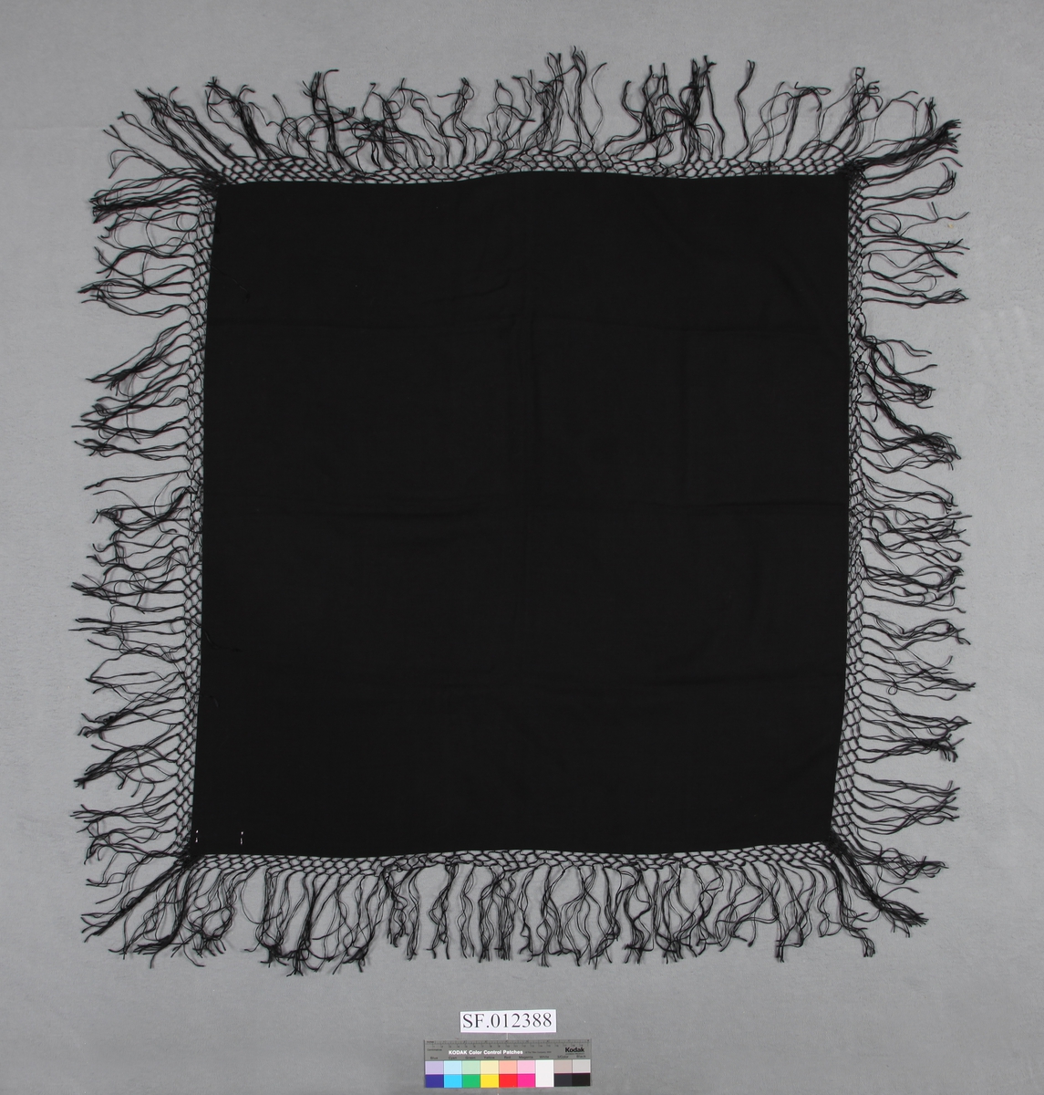 Nesten kvadratisk  forma sjal av svart, vevd ullstoff med frynser på all sidene. Frynsene er ca 3 cm inn mot sjalet, tredde saman og knytta i ruter. Resten, ca 10 cm, heng fritt. . Tre sider er oversydd. Ei side er kanta av stoffet. (Selvage edge)