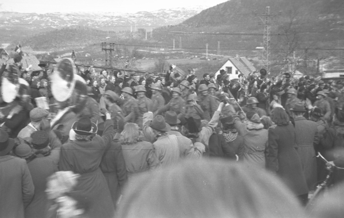 Folkemengde samlet i Stasjonsbakken Narvik Jernbanestasjon. Tuba. Soldater (polititropper?) følger etter korpset. 8. mai 1945
