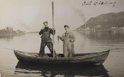 To menn i liten robåt poserer stående i båten med hver sin f