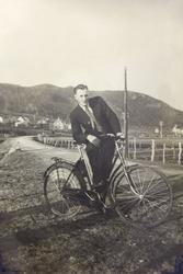 Pent kledd mann med sykkel, fotografert ved en vei. I bakgru