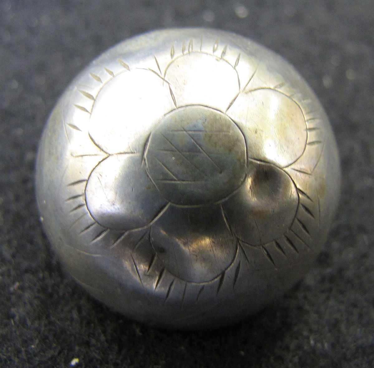 Halvrund, ihålig silverknapp med en enkel ingraverad dekor i form av en blomma. Enligt uppgift ur museets katalog 1904-05 härrör knappen från Norge.