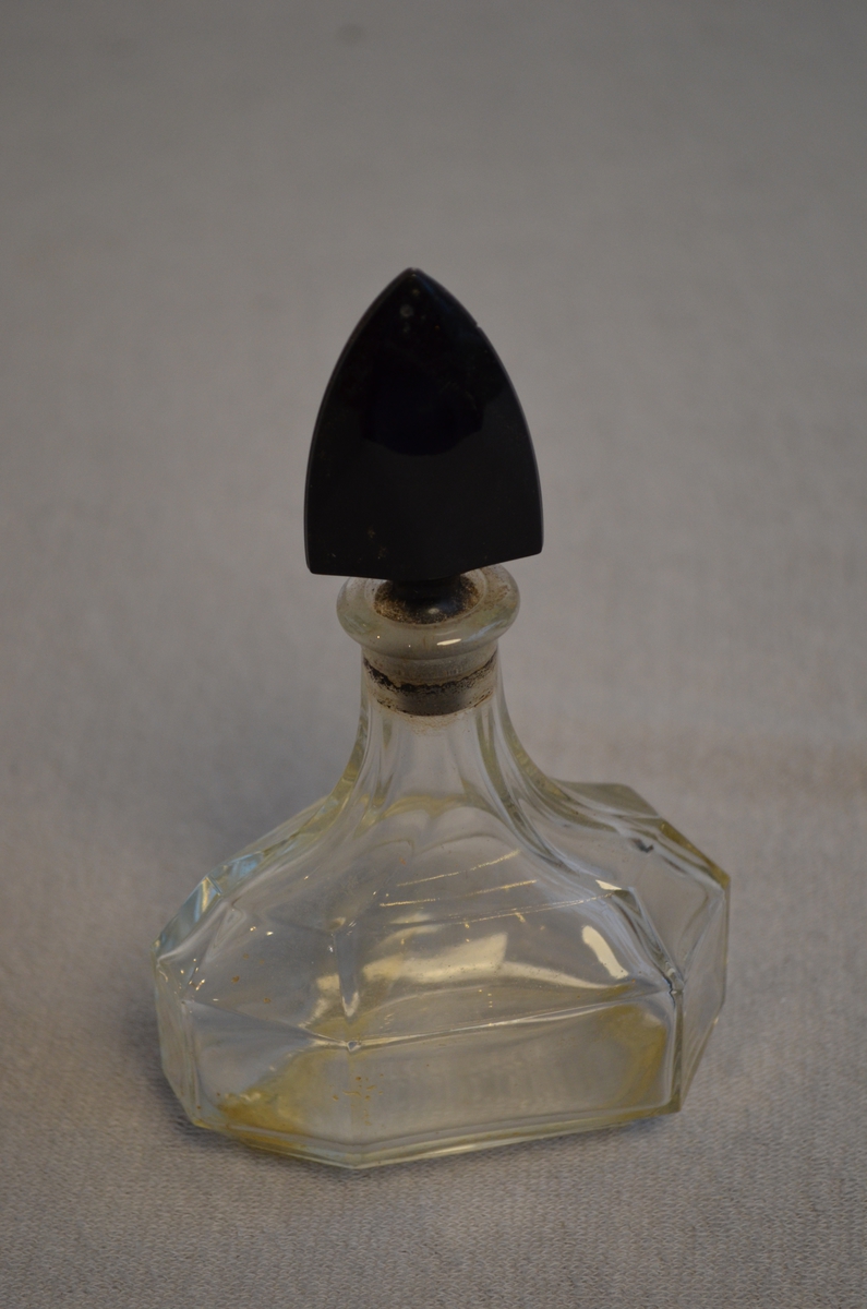 Tom parfymeflaske i gjennomsiktig glas med svart kork, den også i glas. Noko parfyme har tørka i bånn.