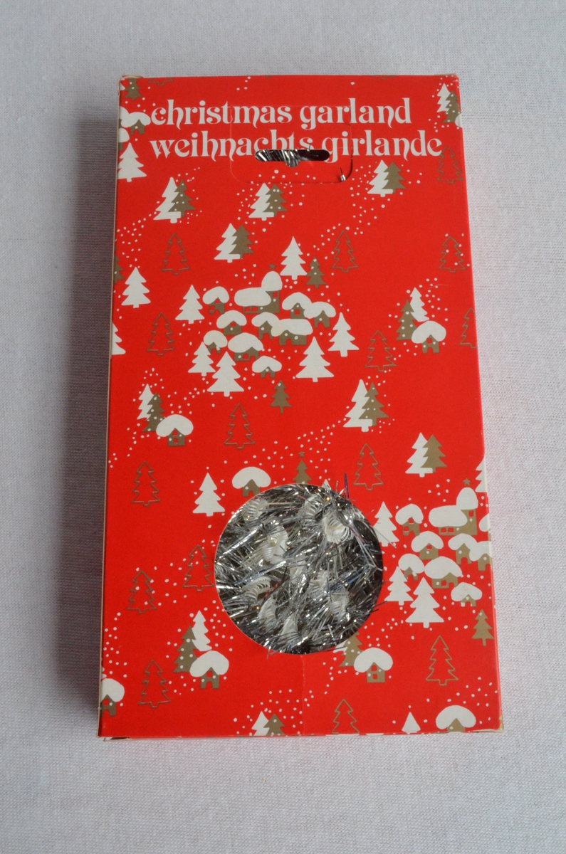 Glitterband i sølv med teksten God Jul. Framleis i embalasjen. Embalasjen er raud og dekorert med juletre og små hus i kvit og gull.