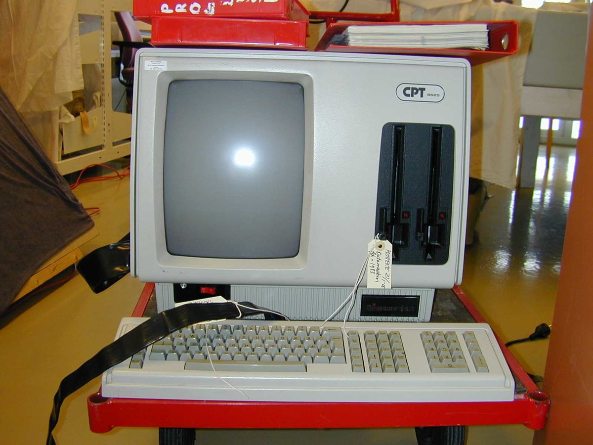 Datamaskin med tastatur og kablar. 
1a= datamaskin der skjerm og maskin er eitt
1b=tastatur
1c=1 raud perm med bruksrettleiing
1d-n=2 raude diskettboksar med 9 store floppydiskettar.