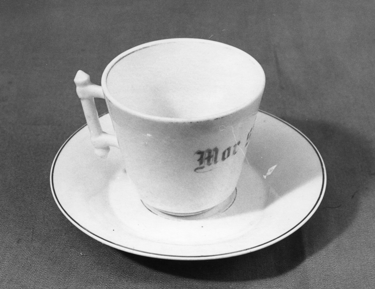 En kopp med tefat i hvitglasert porselen. Koppen har gullstripe nederst og på innsiden øverst. På koppen er det tekst i grått/gullfarge. I den ene siden er det en firkantet hank formet som en stav (stavhank).