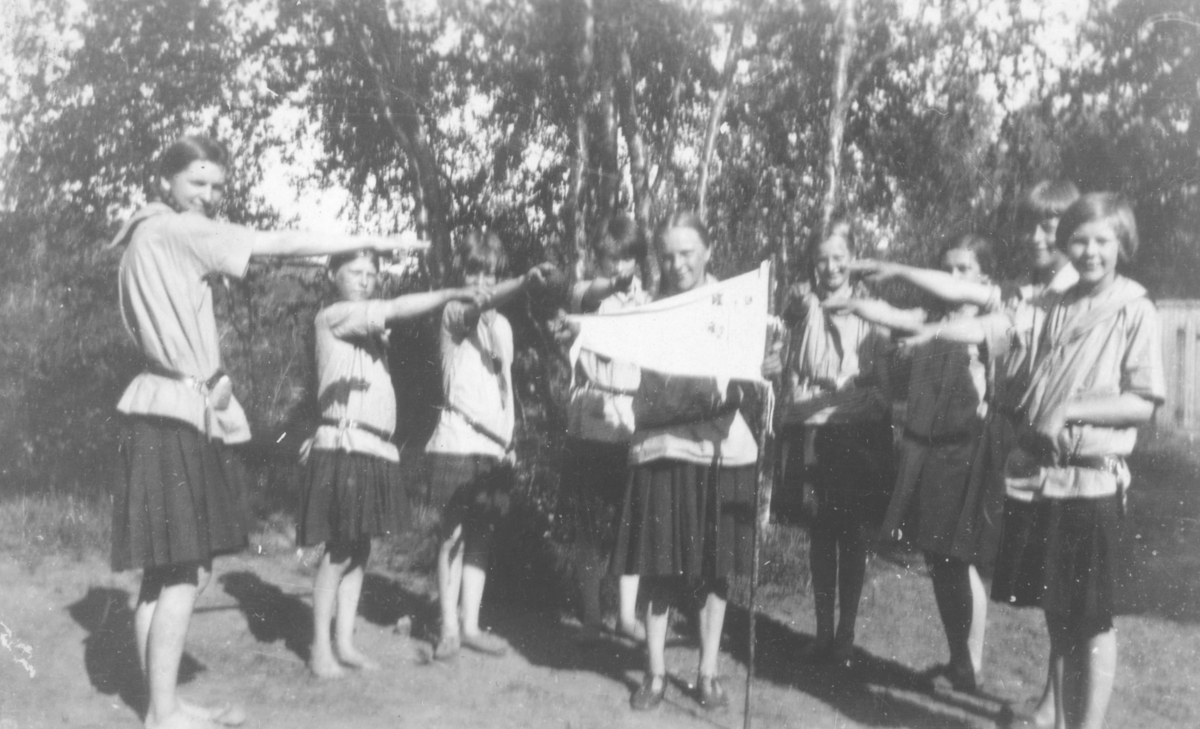 Speiderjenter som gjør flagghilsen. Bildet er tatt i leiren i Rustefjelbma, 1930. Jentene er kledt i speideruniformer, som består av lys skjorte, og mørkt skjørt. I bakgrunnen er det skog