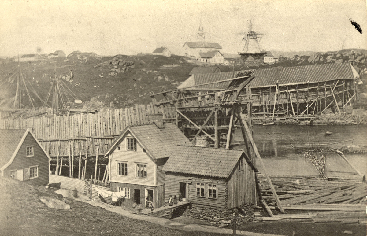 Skipsbygging i Haugeverven på Hasseløy. Tre våningshus og barn  foran i bildet. I bakgrunnen, på fastlandssiden ligger Skåre kirke og en vindmølle.