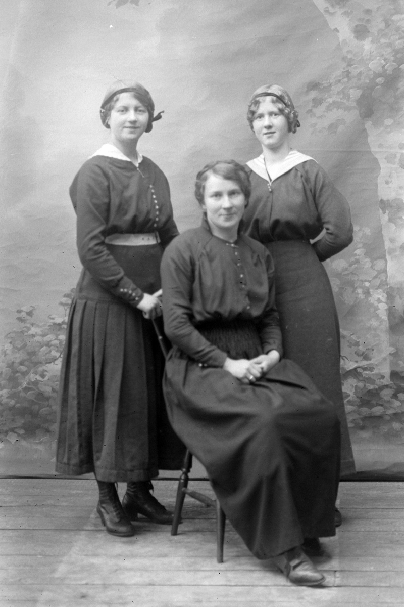 Studioportrett av to kvinner stående bak én som sitter.