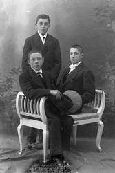 Studioportrett av to menn sittende foran én som står.