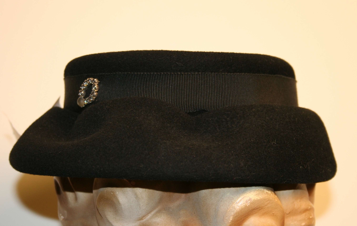 Damehatt i svart filt, med ripsbånd i svart med en liten sløyfe. Rund pull og "diamant" nål som pynt.