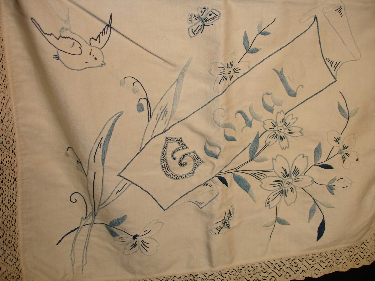 Sengetøypose i hvit bomull med blonderkant rundt, broderi i forskjellige blåfarger, blomstermotiv, fugler, sommerfugler og teksten "Godnat".