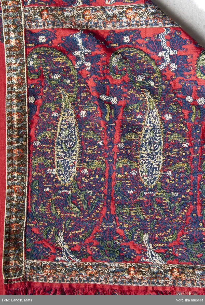 Långschal, europeisk kopia av s.k äkta schal från Kashmir.
Varp och inslag av tvinnat silke, grundbindning; kypert. Handvävd i simpelstol med draget mönster som skyttlats, flotterande trådar på baksidan bortklippta. Röd botten, mönstrade bårder i kortsidorna med 6 stora miribotafigurer i rad. I en ram runt hela schalen en rad med mindre miribota, i färgerna mörkblått, grönt och gult. Smalbård runtom med liten blomranka på vit botten. Längs långsidorna är smalbården vävd separat som ett band och fastsydd mot schalen. Det vita är troligen bomullsgarn och övriga mönsterinslag ullgarn. Fransar i kortsidorna av varpen.
Schalen är typisk för tiden 1815-25. Kan vara vävd i Paisley, jfr. Reilly: The Paisley Shawl, 1996, sid 20.
Har enligt uppgift i huvudliggaren använts som brudschal 1825.
I mycket gott skick.
/Berit Eldvik 2008-08-25