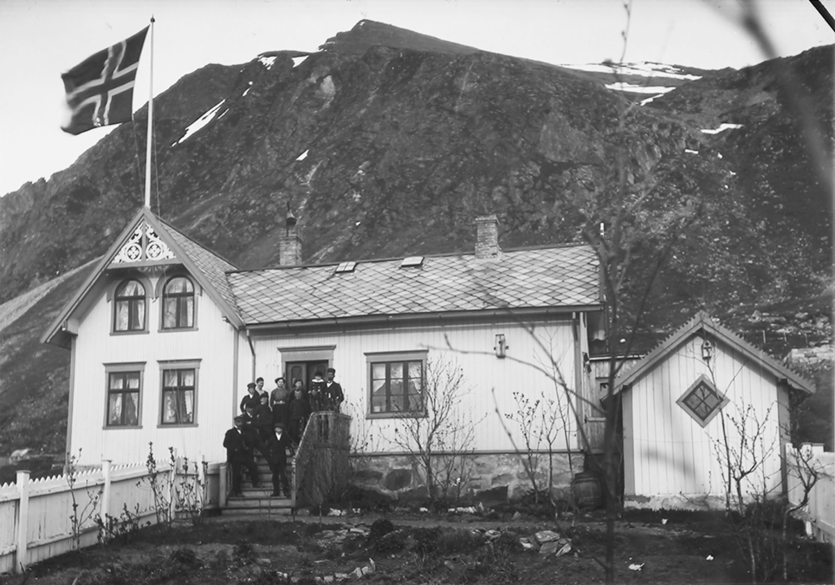 Hus med flaggstang og flagg på taket. Gruppe på trappa. Uthus.Kirkesæter, Ramberg.Her bodde Kristen og Inga Pedersen med familie.