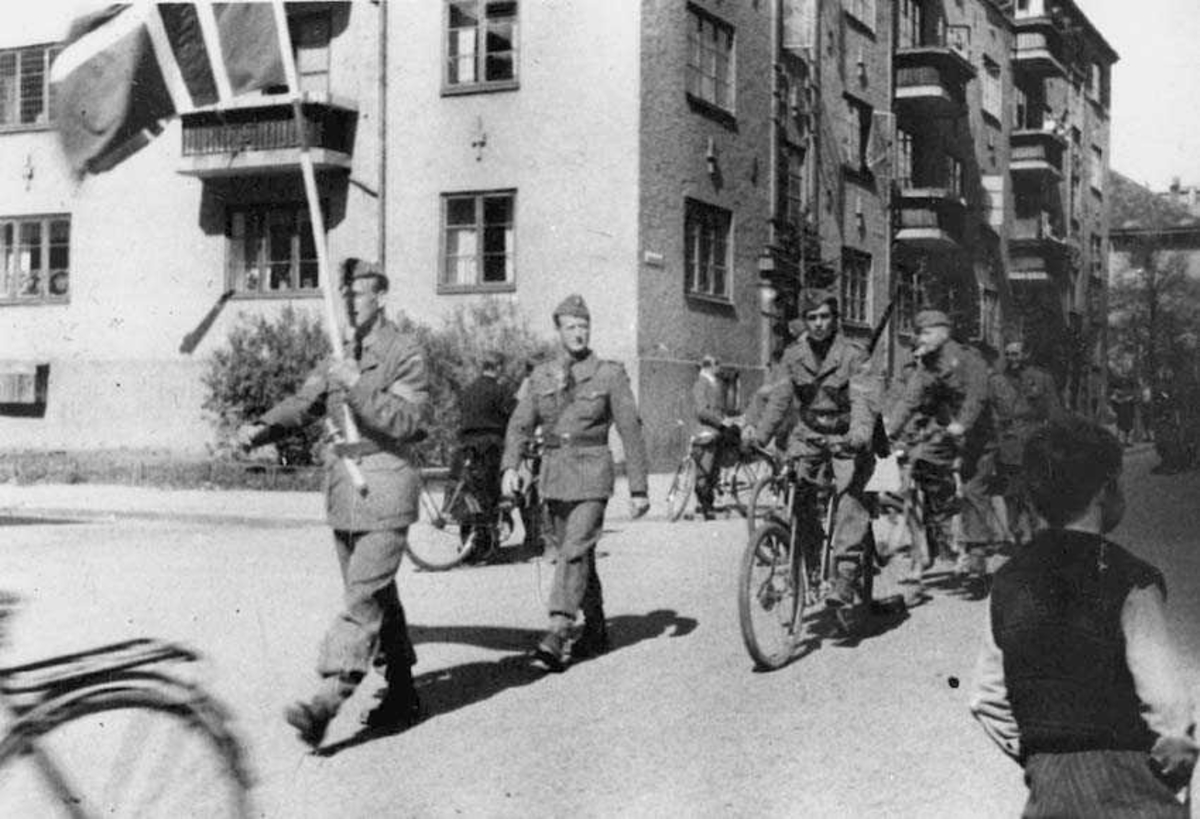 Flere personer i uniform som masjerer.  Den første har et norsk flagg, flere på sykkel. Personer på sidene. Bygning i bakgrunnen.