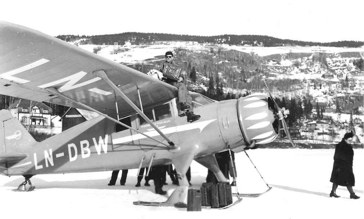 Ett fly på bakken, C-5 Polar  med skiunderstell LN-DBW fra Widerøes Flyveselskap A/S. Flere bensinkanner ved flyet. En person sitter på vingen og fyller bensin på flyet fra en bensinkanne. Flere personer rundt flyet. Snø på bakken.