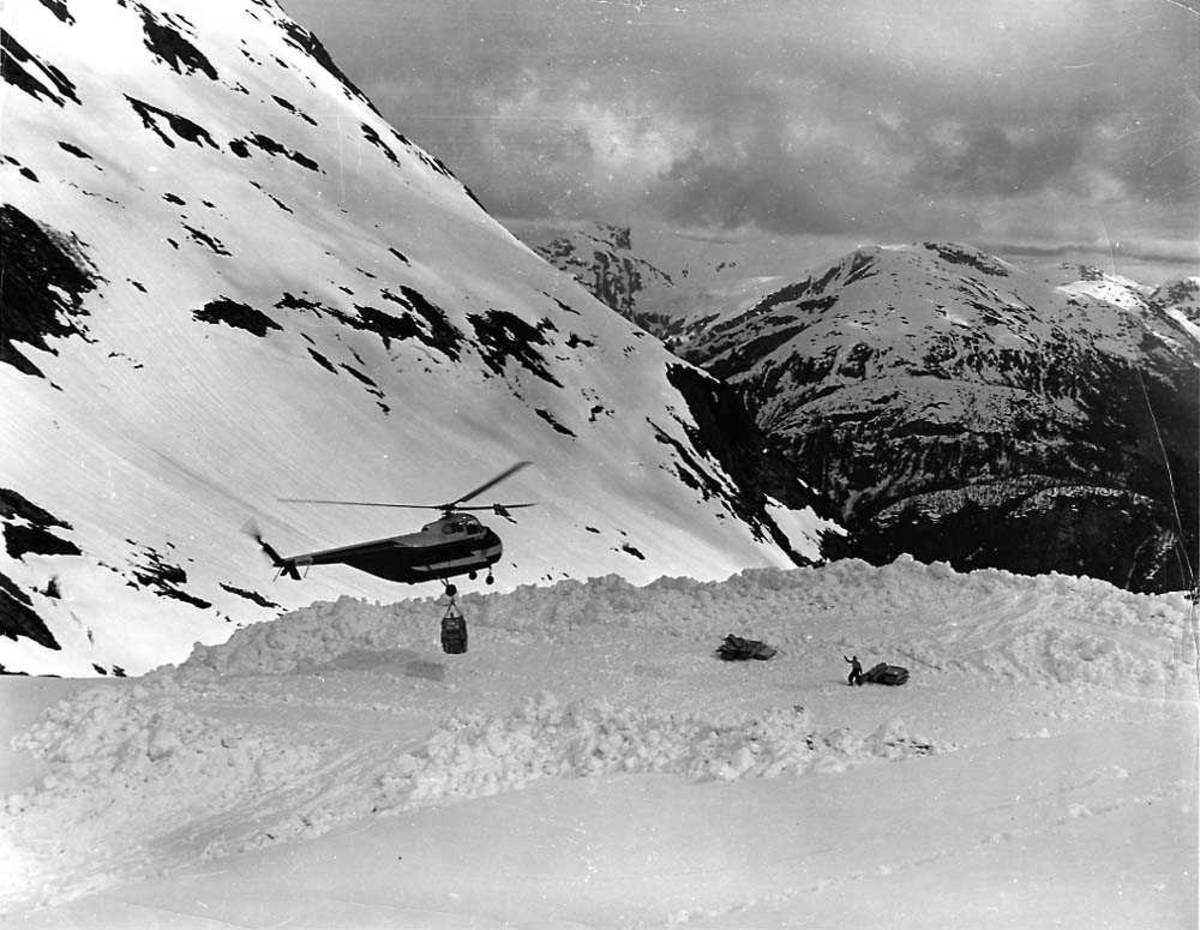 Ett helikopter  i luften med noe last hengende under seg, Sikorsky S-55 H-19 Chickasaw. Fjell i bakgrunnen. Snø på bakken.