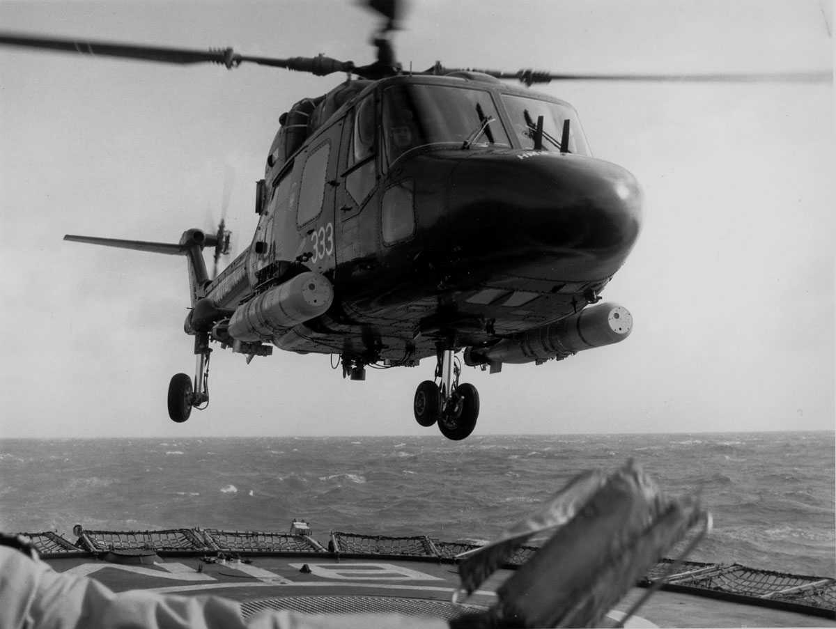 Ett helikopter i luften over helipad/hoveddekk på skip. Westland Lynx , sidedør merket 333. To torpedoer, en på hver side av helikopteret.