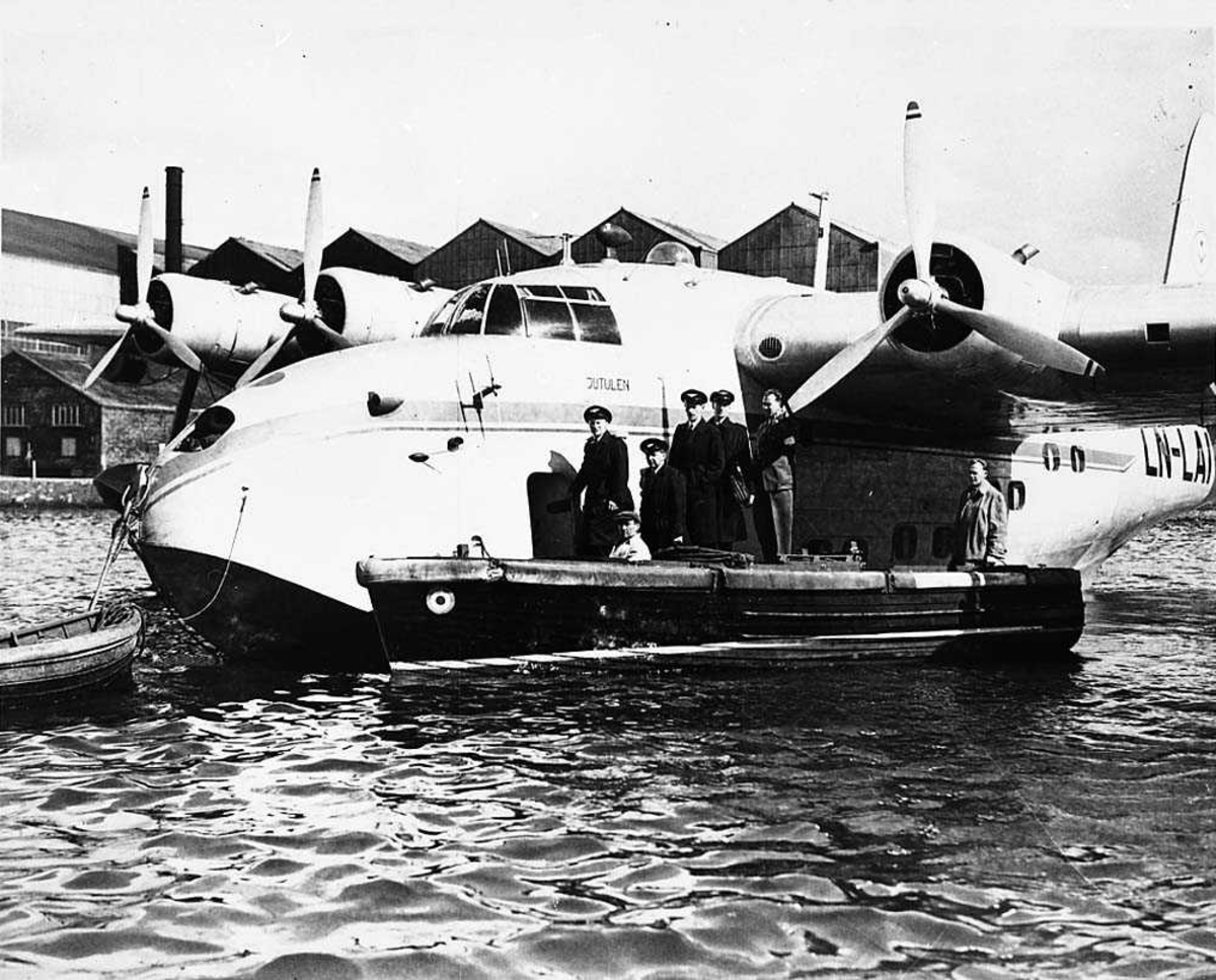 Sjøflyhavn. Ett sjøfly som ligger fortøyd på vannet, Shorts S.25 Sandringham LN-LAI "Jutulen" fra DNL. En båt ligger ved flyet med flere personer ombod, bl.a. flymannskap.