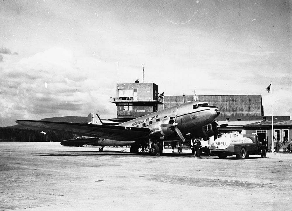 Lufthavn. Ett fly på bakken som blir tanket opp, Douglas DC-3/C-47 Dakota fra Norwegian Air Lines (DNL). En tankbil foran flyet. Bygninger i bakgrunnen.