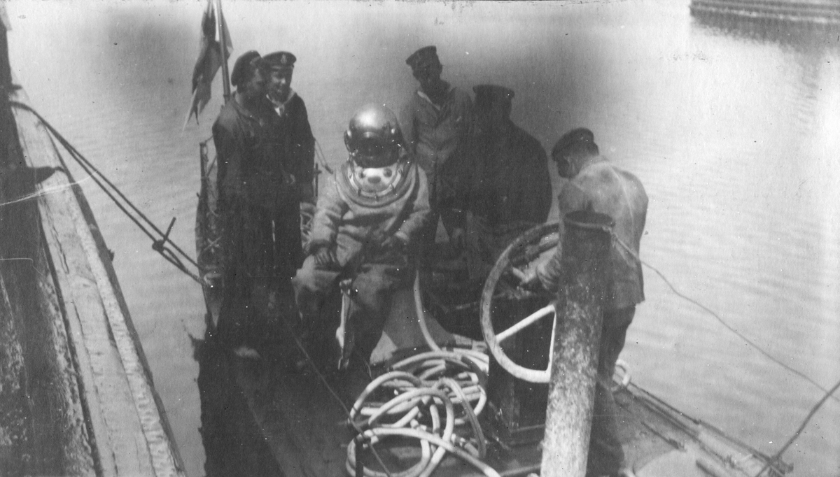 Fem män på båt förbereder en sjätte för nedstigning, iklädd dykardräkt.