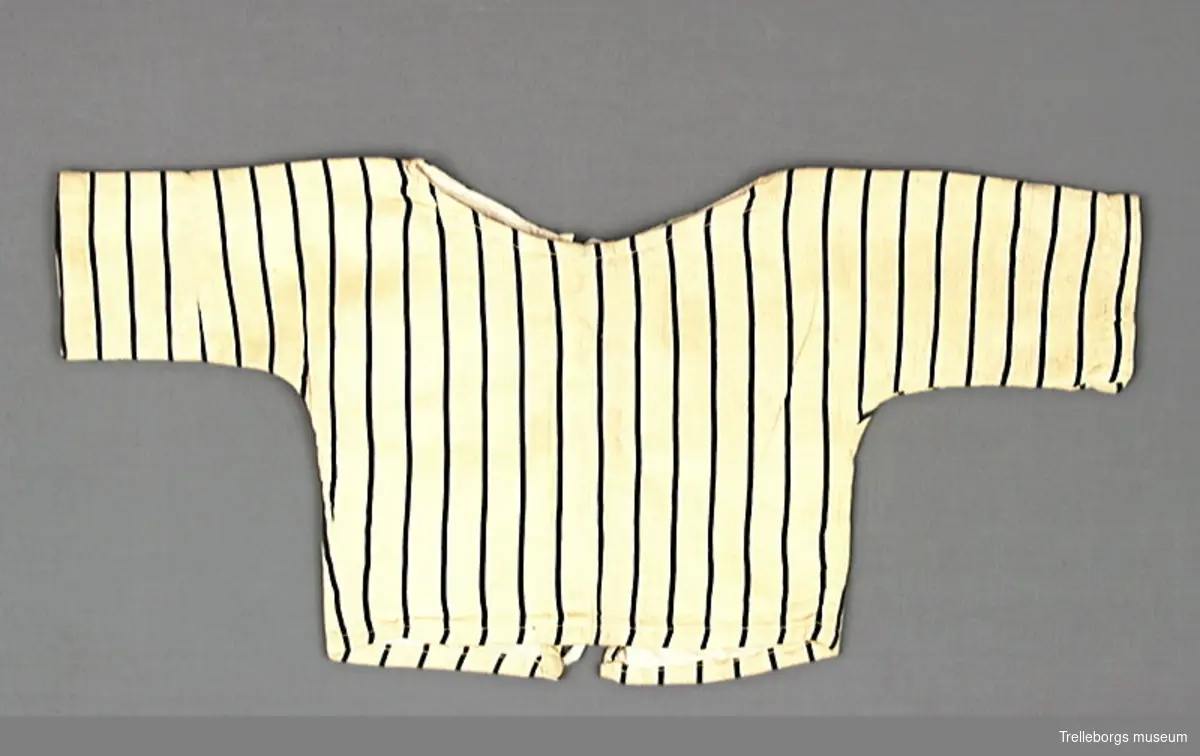 Babyskjorta med långa ärmar i ett gult tyg med svarta ränder. Skuren i ett stycke så det finns inga axel eller ärmsömmar. Halsringningen är rund och helt öppen framtill, den knyts ihop med två par bomullsband. Hela skjortan är fodrad med ett vitt bomullstyg.