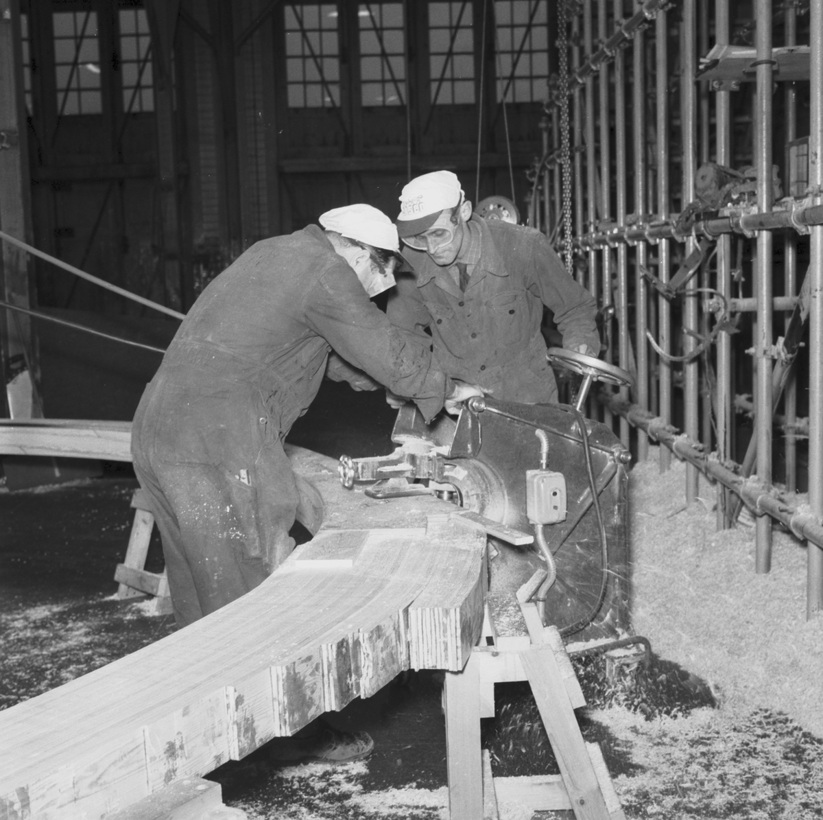 Bilden visar två varvsarbetare som bearbetar en limad träspant för träminsveparen M 60 med en maskin.
