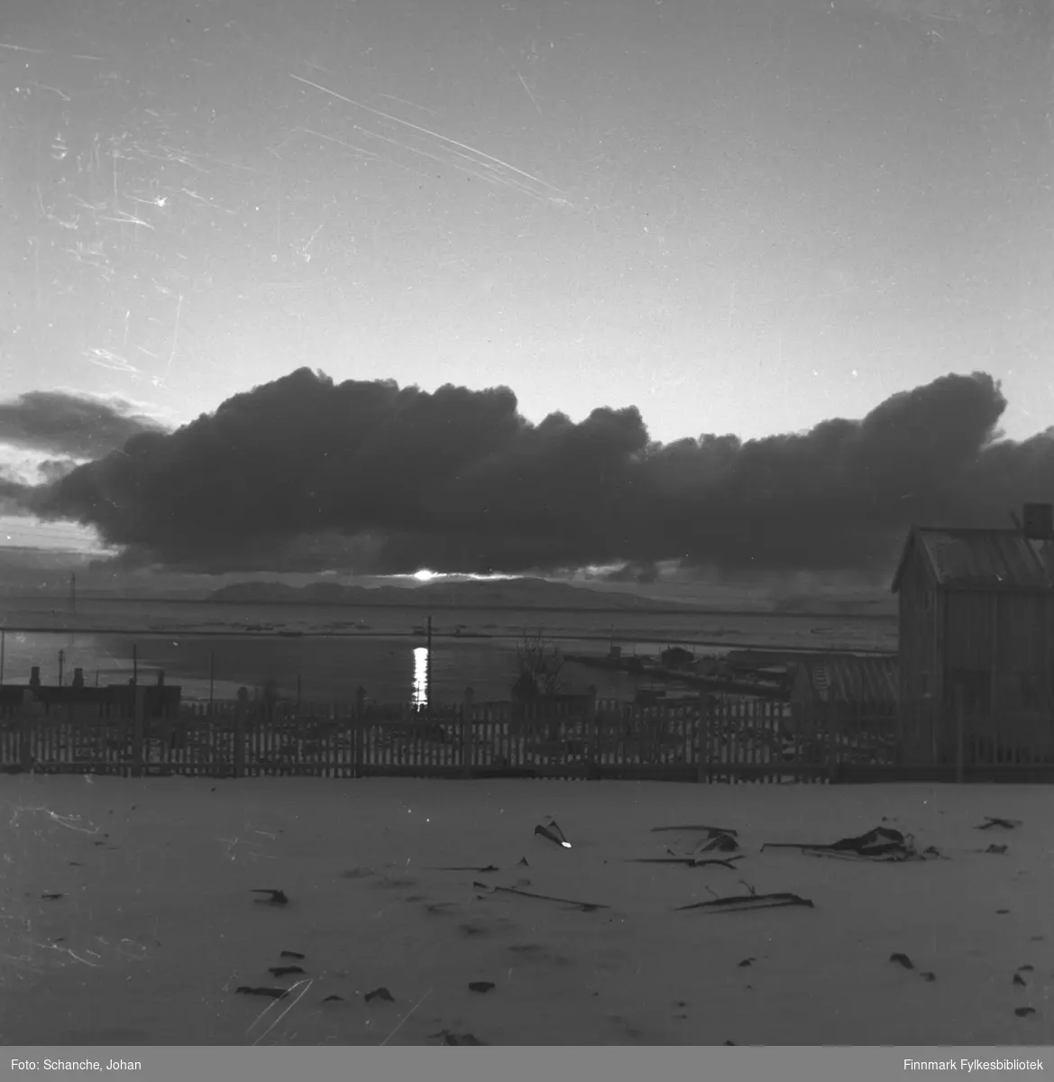 Solnedgang fotografert i Vadsø våren -46. Foran på bildet  et nybyggdt hus med gjerde i Indrebyen, bak den en del av Midtbyen: byggninger og fjorden.  Solen går ned bak Øya og speiler seg i havet.
