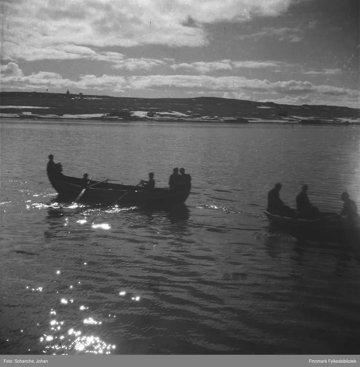 To robåter ligger i Varangerfjorden utenfør Vadsø -46.  To menn ror mot land. Båtene er fotografert mot sol slik at både båtene og menneskene om bord blir mørke siluetter i havet.  Det er litt snø på fjellet.