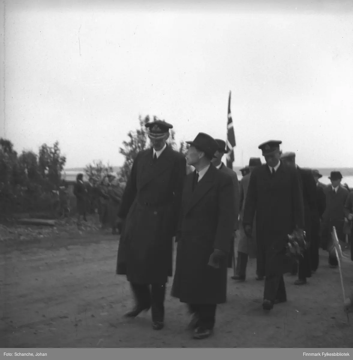 Kong Haakons besøk i Vadsø i 1946. Kong Haakon VII, fylkesmann Hans Gabrielsen og adjutant kommandørkaptein Bruusgaard  (med blomsterbukett i hand) går langs gata i Vadsø. Folkemengde på vegkanten.