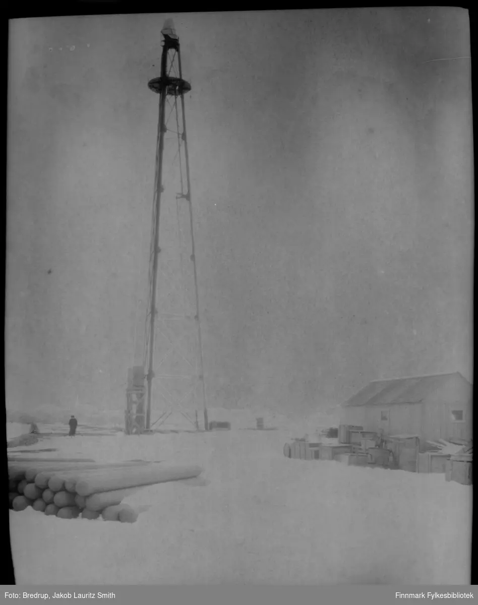 Kvinne fotografert ved luftskipsmasta på Vadsøya, det er full vinter på bildet.  Det ligger stablet gassflasker, tønner og esker ved masta og ved hytta som ble bygd i tilknytning til masta.  Antakelig forberedes ankomsten til ett av luftskipene i mai samme år, enten bildet er tatt i 1926 eller 1928.