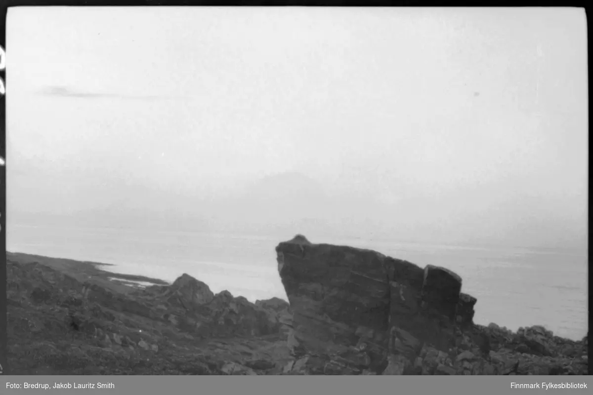 Bildet er fotografert fra en høyde ned mot havet.  Landskapet er fullt av steiner og klipper, midt i bildet dominerer en stor stein.  Bildet kan være tatt mange steder i Finnmark.